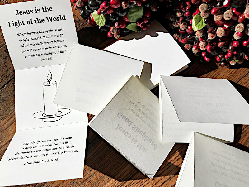Christmas Names of Jesus Cards for Kids folded near red and green decorations. "Jesus is the light of the world" tri-fold card is open with picture of candle, Bible verse, and explanation.