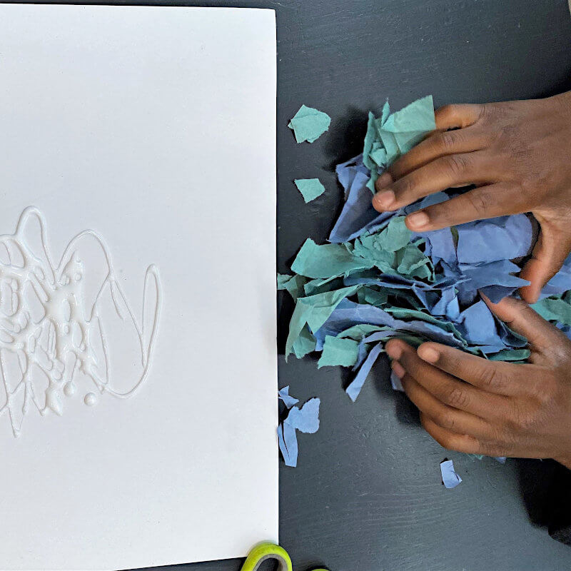 Child's hands gathering torn green and blue papers next to a white paper with glue as he prepares to make an art project for the book Perfect Square