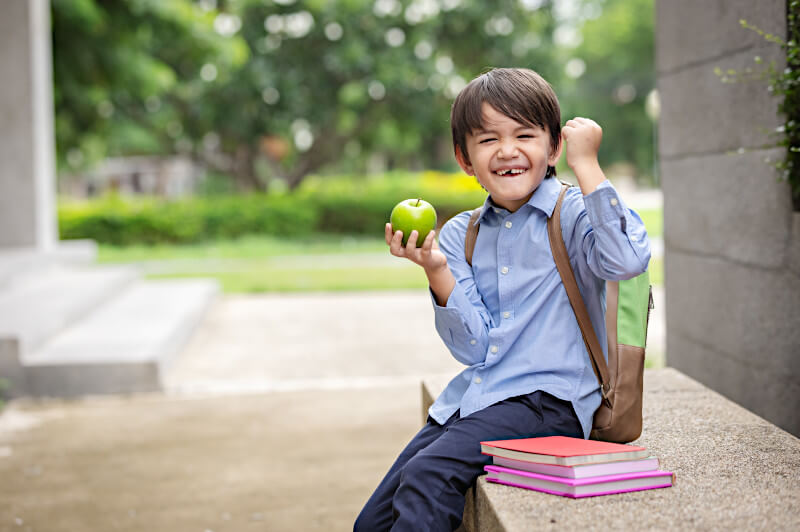 A little boy with a backpack and green apple sits on a bench near a stack of book with a happy, confident, excited expression meant to convey a love for these back-to-school books for Christian families