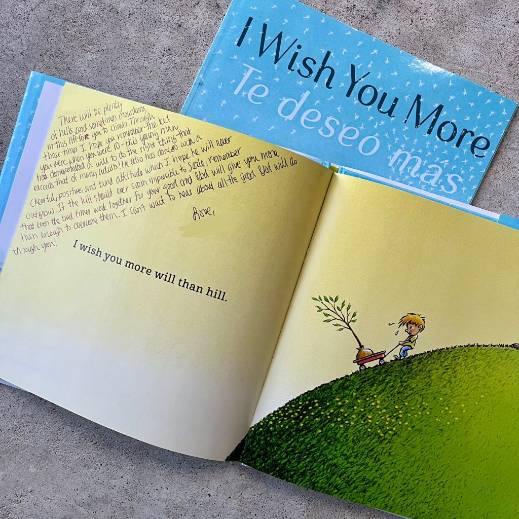 An opened copy of I Wish You More signed by a teacher as a keepsake gift for a child. It's open to the page "I wish you more will than hill" where an illustration shows a boy pulling a tree up a hill in a wagon.