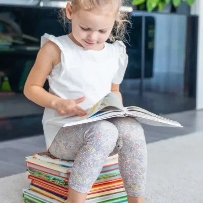 Child is reading a book while sitting on a stack of books