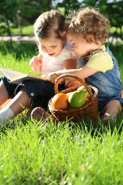 Two young children reading an Easter book outside in the grass with a basket of goodies nearby
