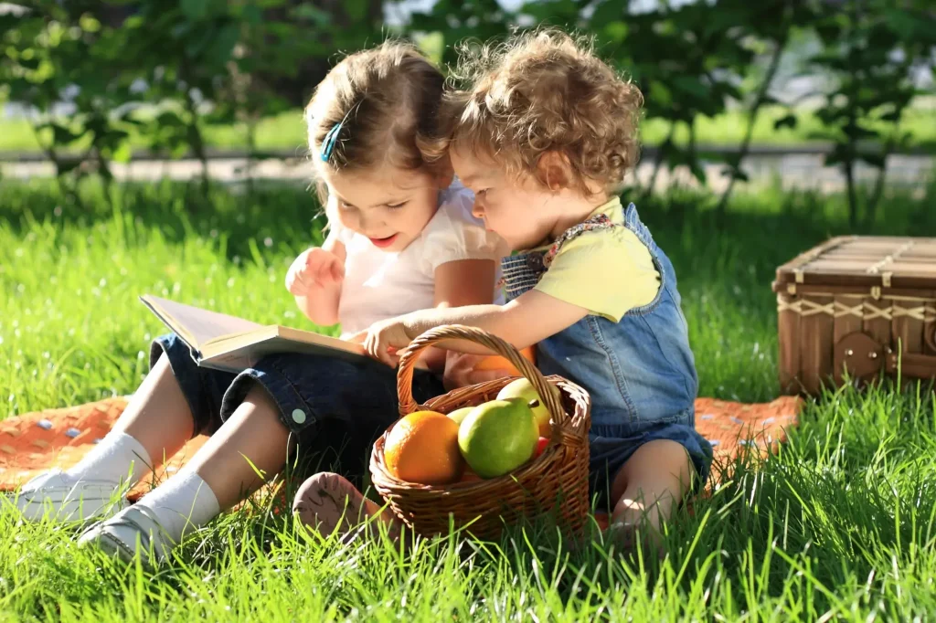 Two young children reading an Easter book outside in the grass with a basket of goodies nearby